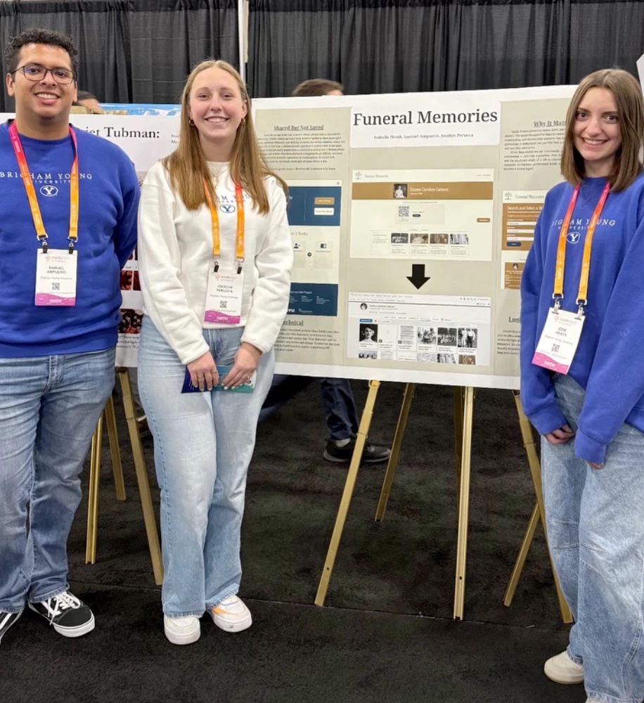 Three students in BYU sweatshirts and RootsTech lanyards standing beside a printed Funeral Memories poster on an easel